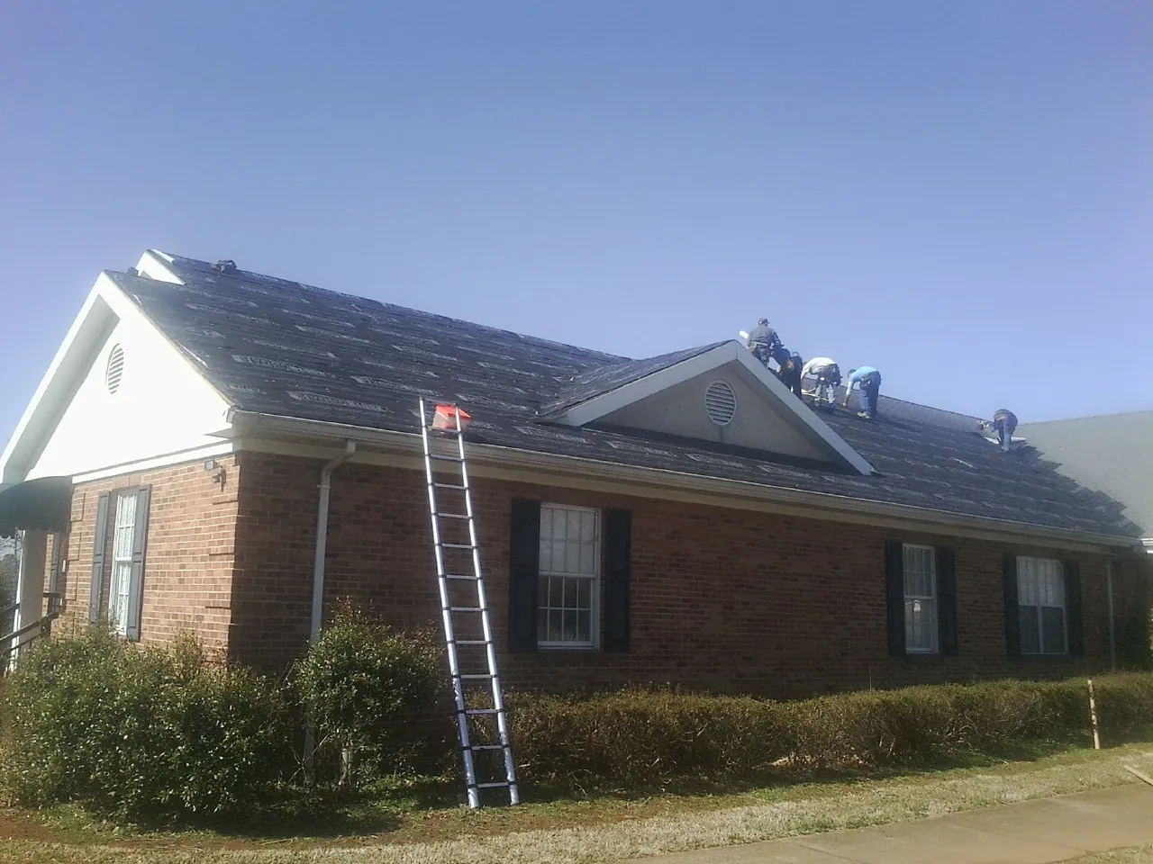 Crew tearing shingles off a brick ranch roof with a ladder set — Metal Roof Repair work by our crew in Cantonment