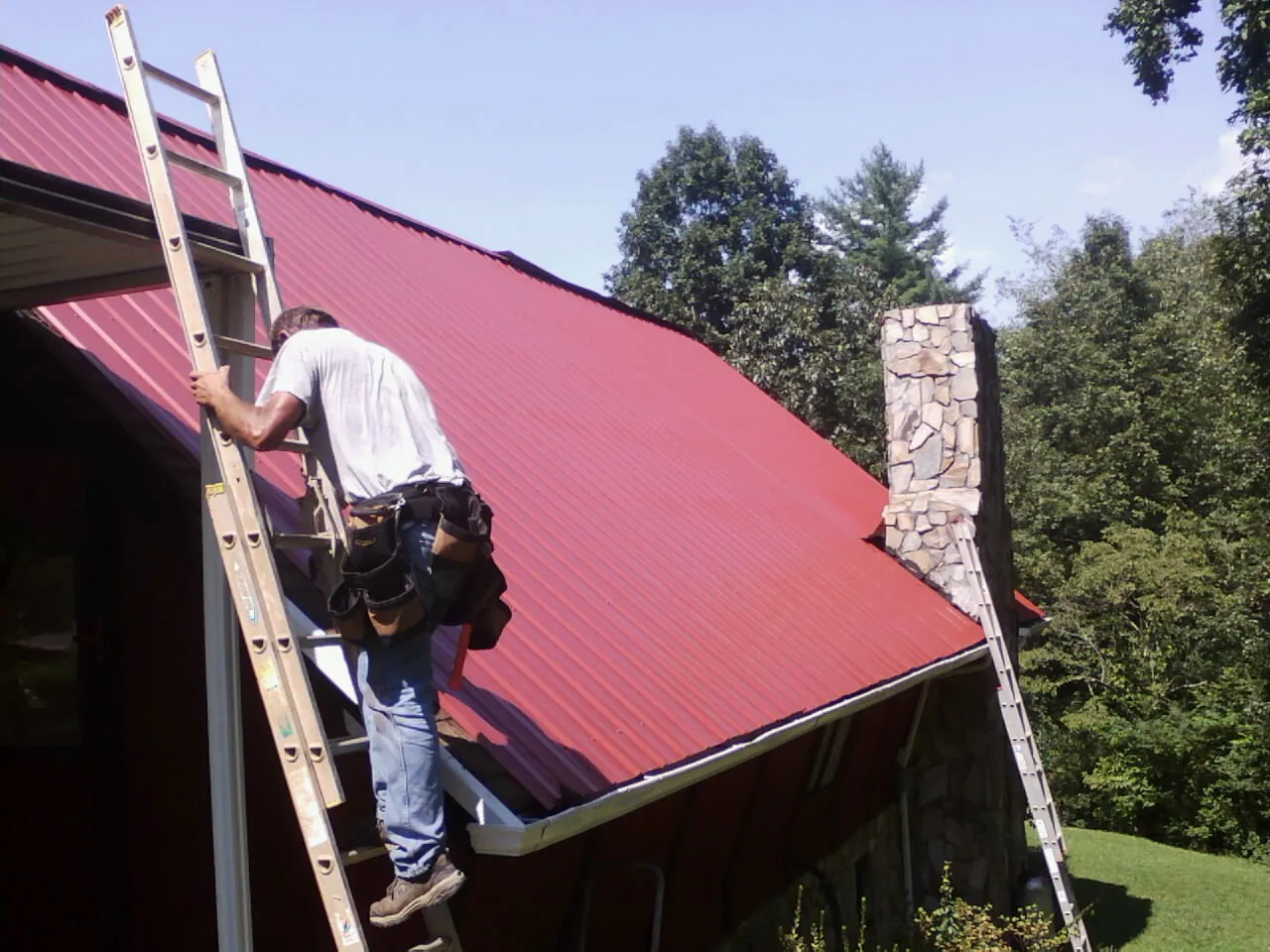 Roofer on a ladder beside a red metal roof and stone chimney — Soffit & Fascia Repair by Roofing Brothers in Cantonment